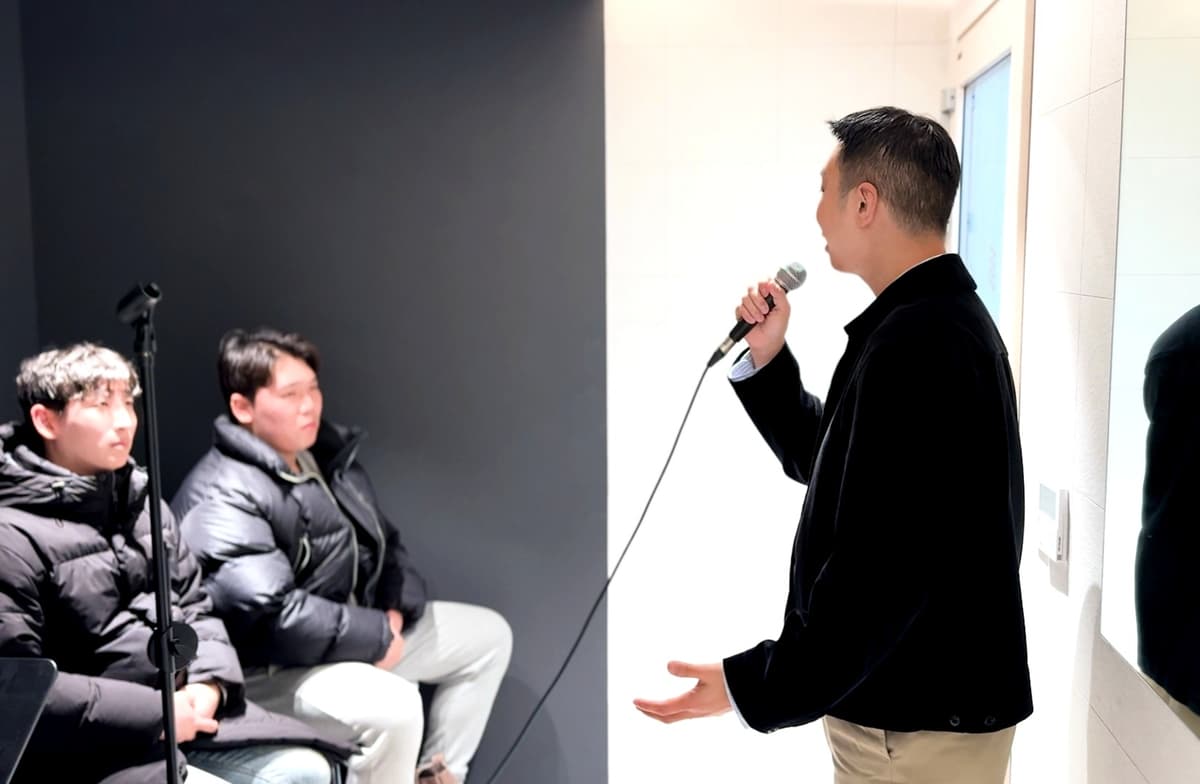 마이크 든 남성 강사가 두 학생에게 보컬 레슨을 진행하고 학생들이 경청한다.
