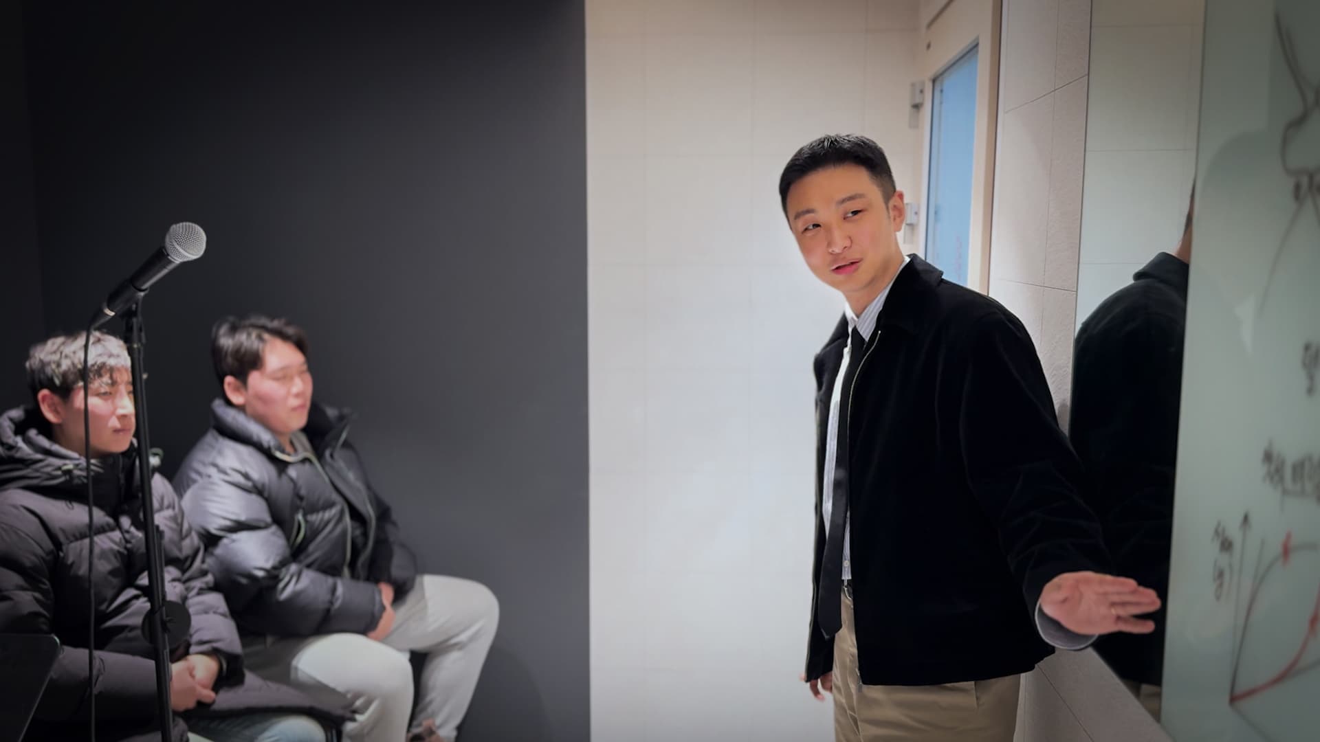 강사가 마이크 옆에 앉은 두 학생에게 화이트보드를 가리키며 설명한다.