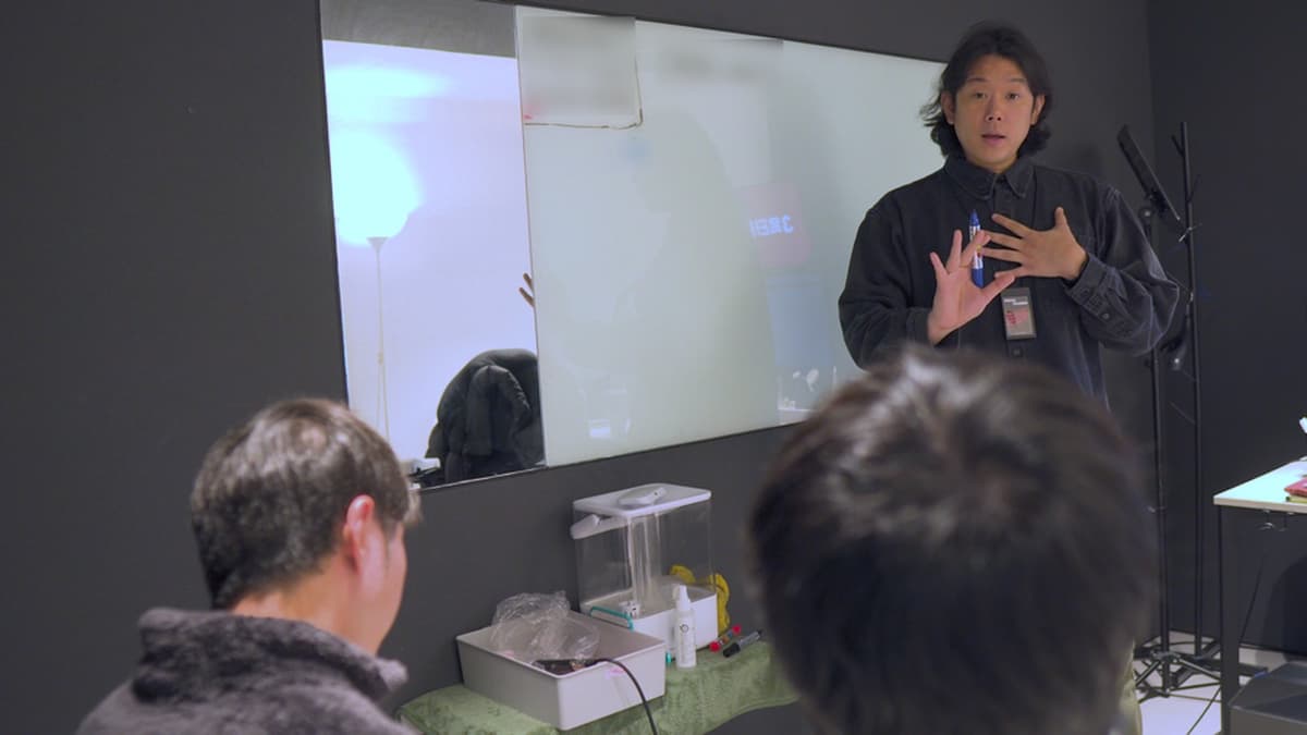 보컬 강사가 학생들 앞에서 펜을 들고 노래 자세를 설명한다.