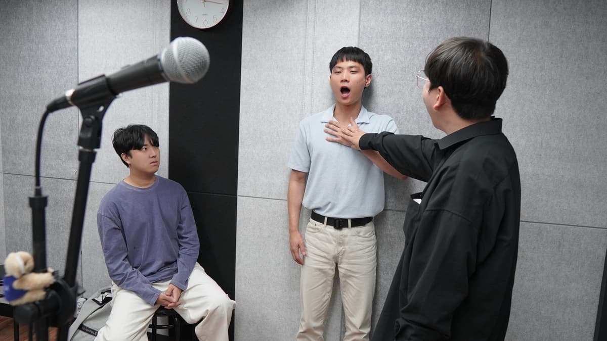 보컬 트레이너가 학생의 발성 위치를 잡고, 다른 학생은 마이크 앞에서 수업을 지켜본다.